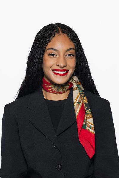 Woman with braided hair, red lipstick, black coat, and colorful scarf smiling.