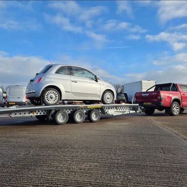 A red pickup truck towing a silver car on a trailer in a parking lot.