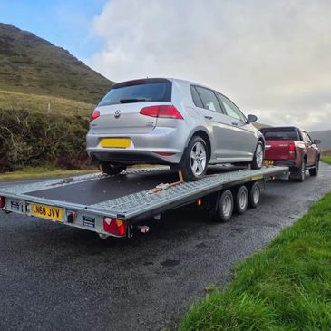 Silver Volkswagen car loaded on a trailer towed by a red pickup truck.