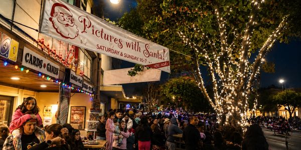 Crowd gathered for Pictures with Santa event at night under decorated tree.