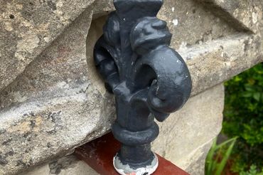 Close-up of a decorative black iron finial on a rusty red metal bar against a stone wall.