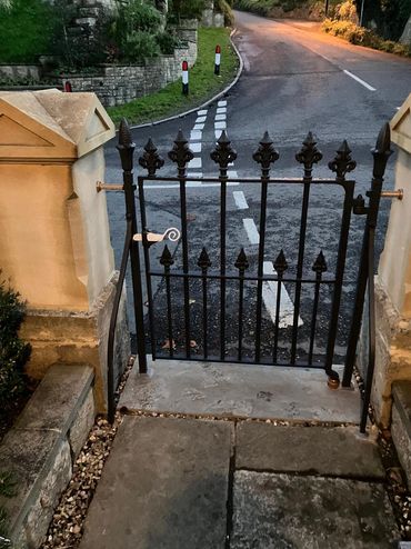 Black wrought iron gate at the entrance to a stone pathway beside a road.