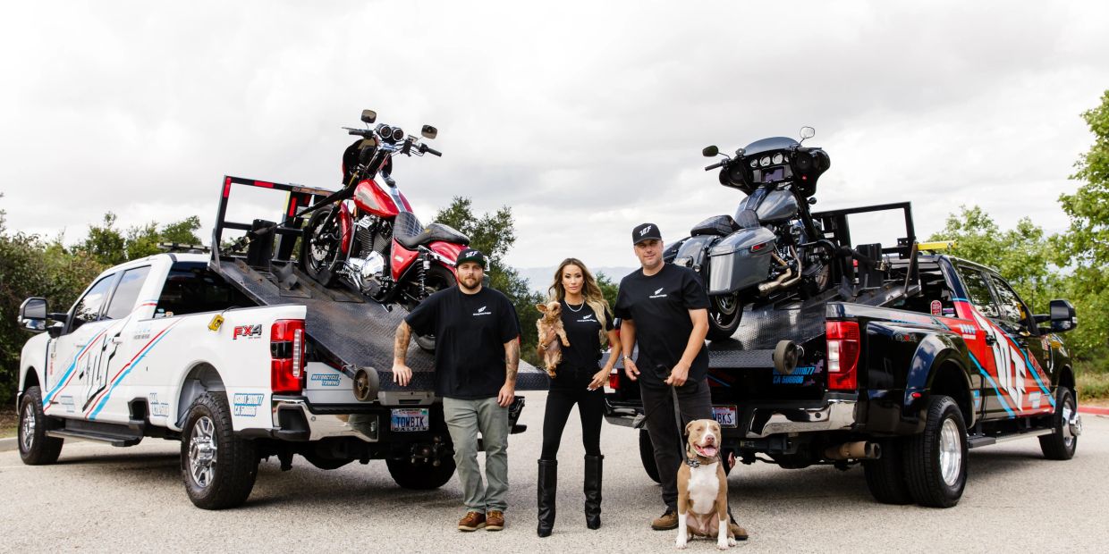 Three people with dogs between two trucks carrying motorcycles.