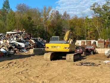 Yellow excavator at a scrapyard with piled metal and damaged cars.