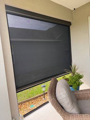 Outdoor wicker chair with cushion beside a retractable black screen and potted plants.