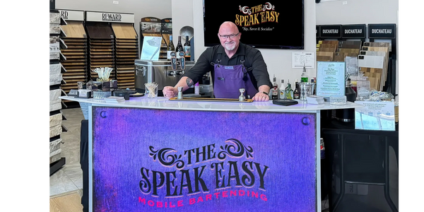 A bartender stands behind a mobile bartending counter with a purple "The Speak Easy" sign.