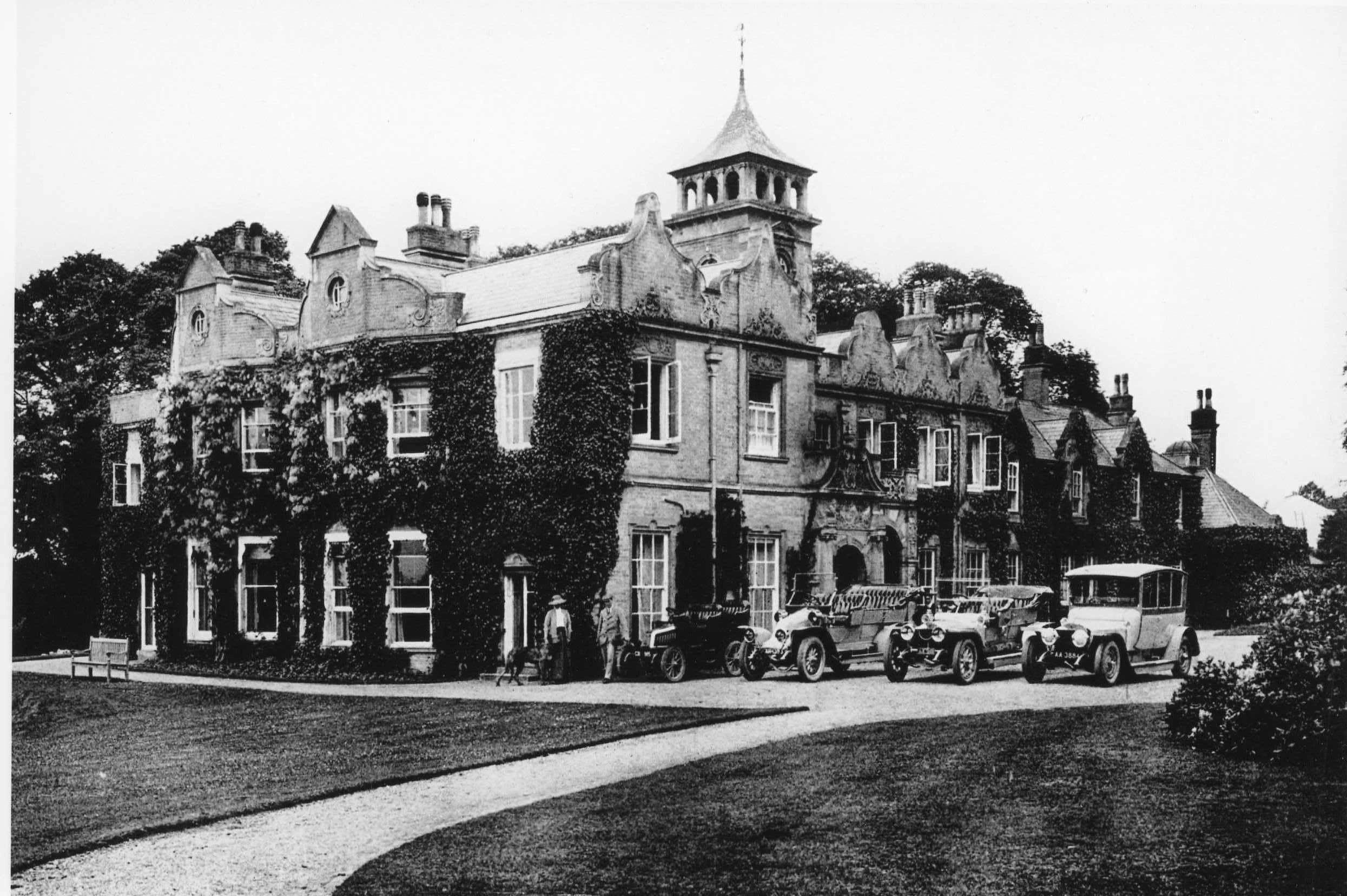 Vintage black and white photo of a large ivy-covered mansion with early 20th-century cars parked outside.