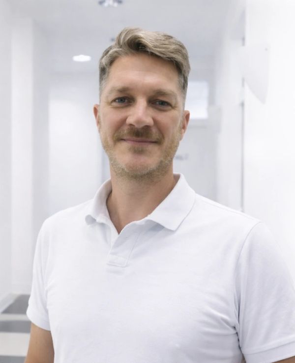 Smiling man in a white polo shirt stands in a bright, modern hallway.