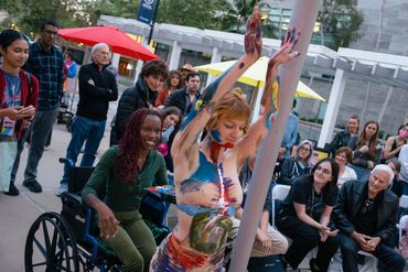 Casey presents their body to a smiling black woman in a blue wheelchair with a pallet of paint