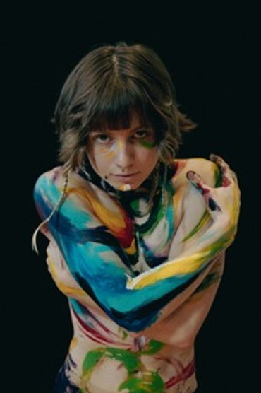 Painted with bright colors flowing into one another, Casey hugs themself tight and stares intently