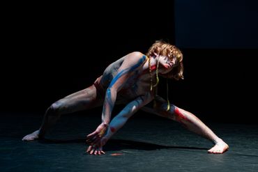 Leaning in a wide stance with streaks of blue and red paint, Casey drags their hands on the ground