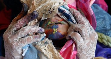 lacy gloved hands peel back layered fabric to expose a mouth covered in multi-colored paint.