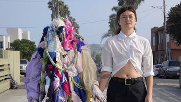A shroud of colorful clothes with no face stands holding hands with a person in a white button down