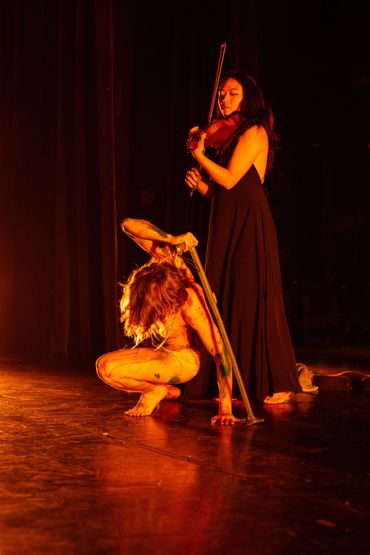 Violinist in black dress looms over Casey as they crouch propped up by a cane