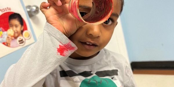 A young boy looks through a painted cardboard tube, wearing a dinosaur shirt.