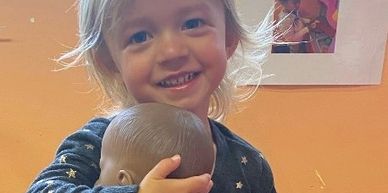 Smiling toddler holding a baby doll in a playful classroom setting.