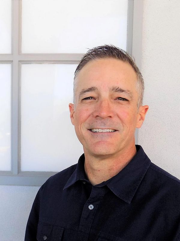 Smiling man in a dark shirt stands by a window with frosted glass panes.