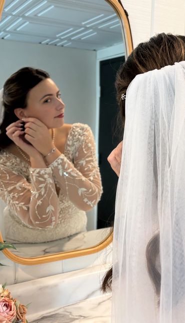 Bride adjusting earrings while gazing in the mirror, wearing a lace wedding dress and veil.