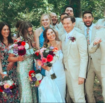 Joyful wedding group with bride, groom, and friends outdoors.