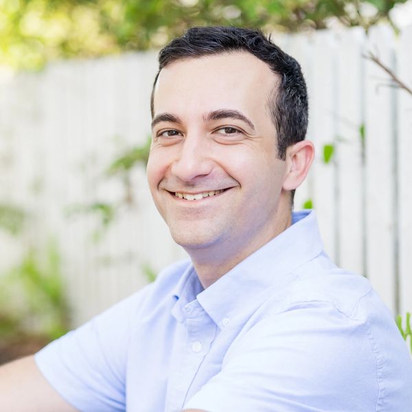 Smiling man in light blue shirt outdoors with a blurred fence background.