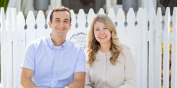 Smiling couple sitting in front of a white picket fence on a sunny day.