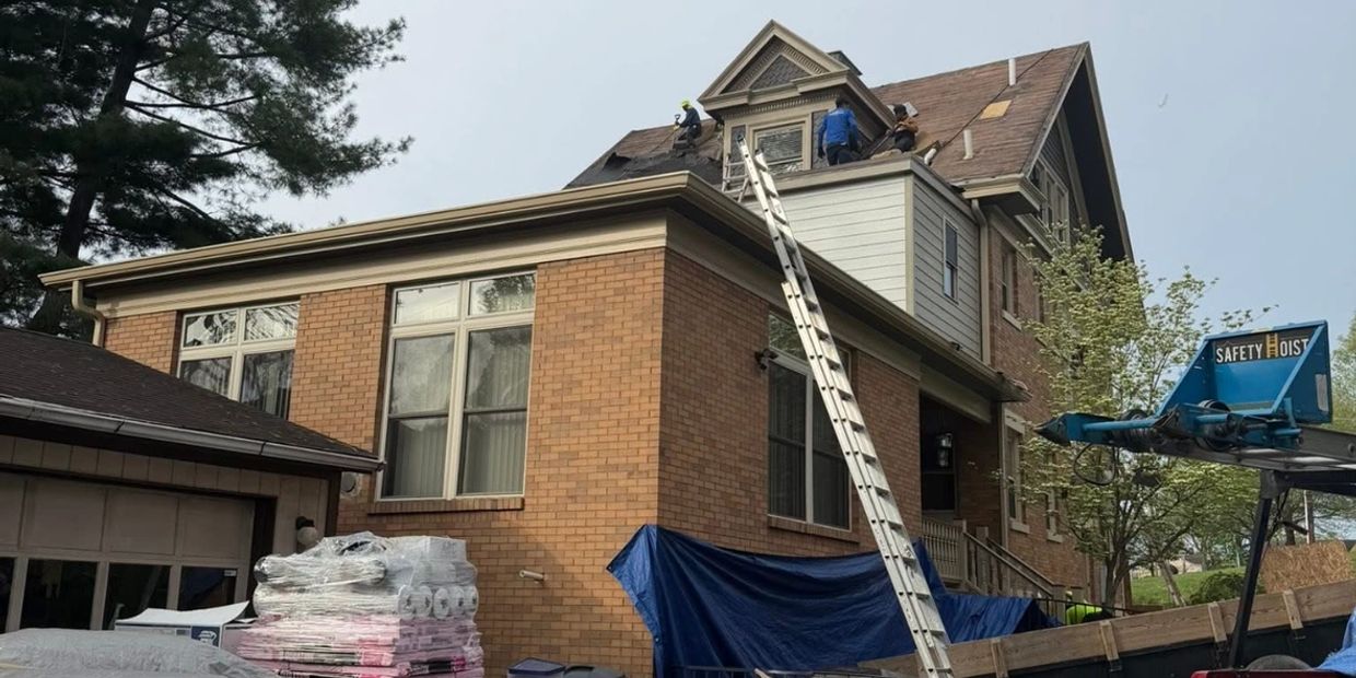 Workers repairing the roof of a two-story brick house using ladders and safety equipment.