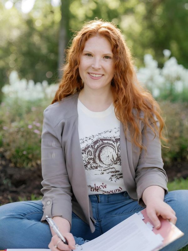 A smiling woman with red hair sitting cross-legged outdoors, holding a pen and papers.