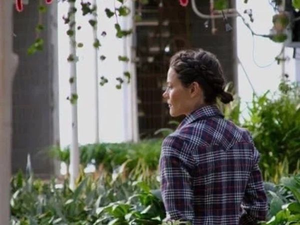 Woman with braided hair in a plaid shirt amidst greenery.