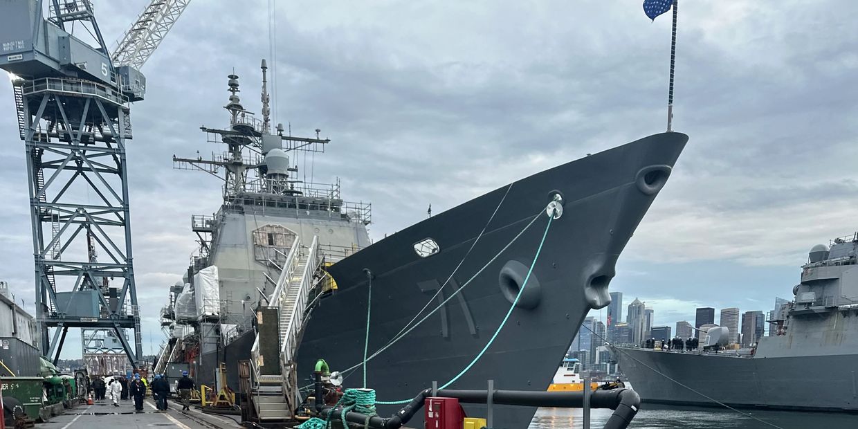 A large naval ship docked at a harbor with workers nearby under a cloudy sky.