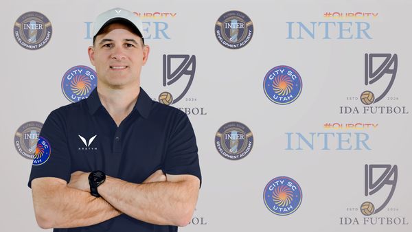 Man in navy polo and cap poses with arms crossed in front of sports logos.