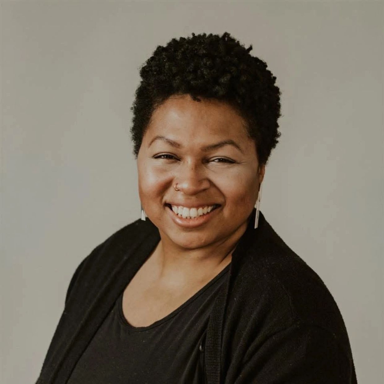 Smiling woman with short curly hair and nose ring against a neutral background.