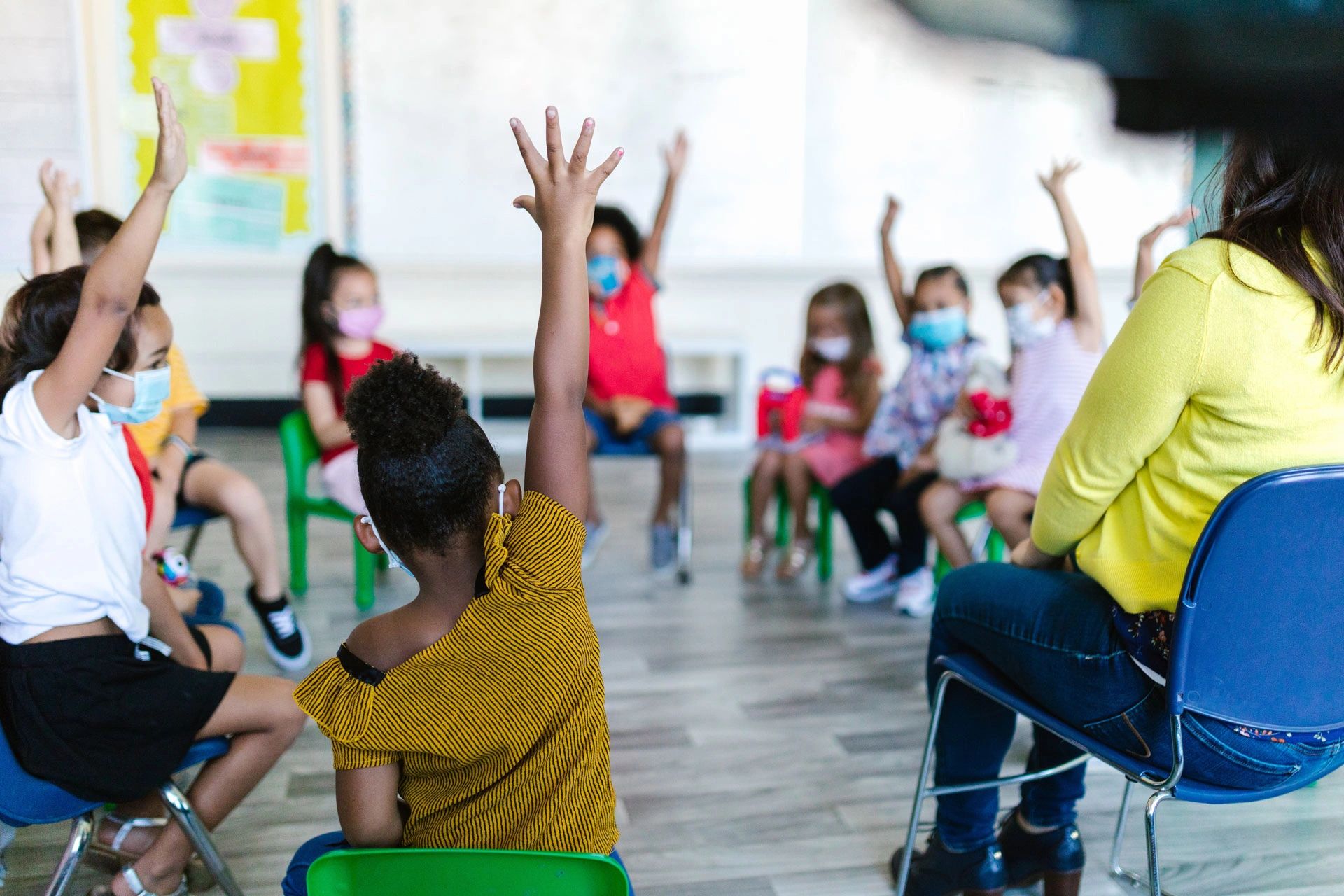 Young children sitting in a circle, many are raising their hands to answer a question.