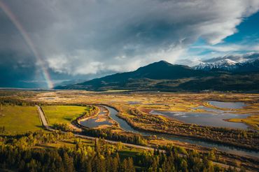 A vibrant rainbow arcs over a lush valley with winding rivers and snowy mountains.