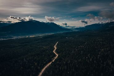 A winding road through dense forest leading to distant mountains under a cloudy sky.
