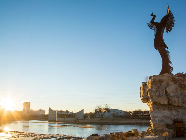 Silhouette of a metal statue on a rock at sunset by a river with city buildings in the background.