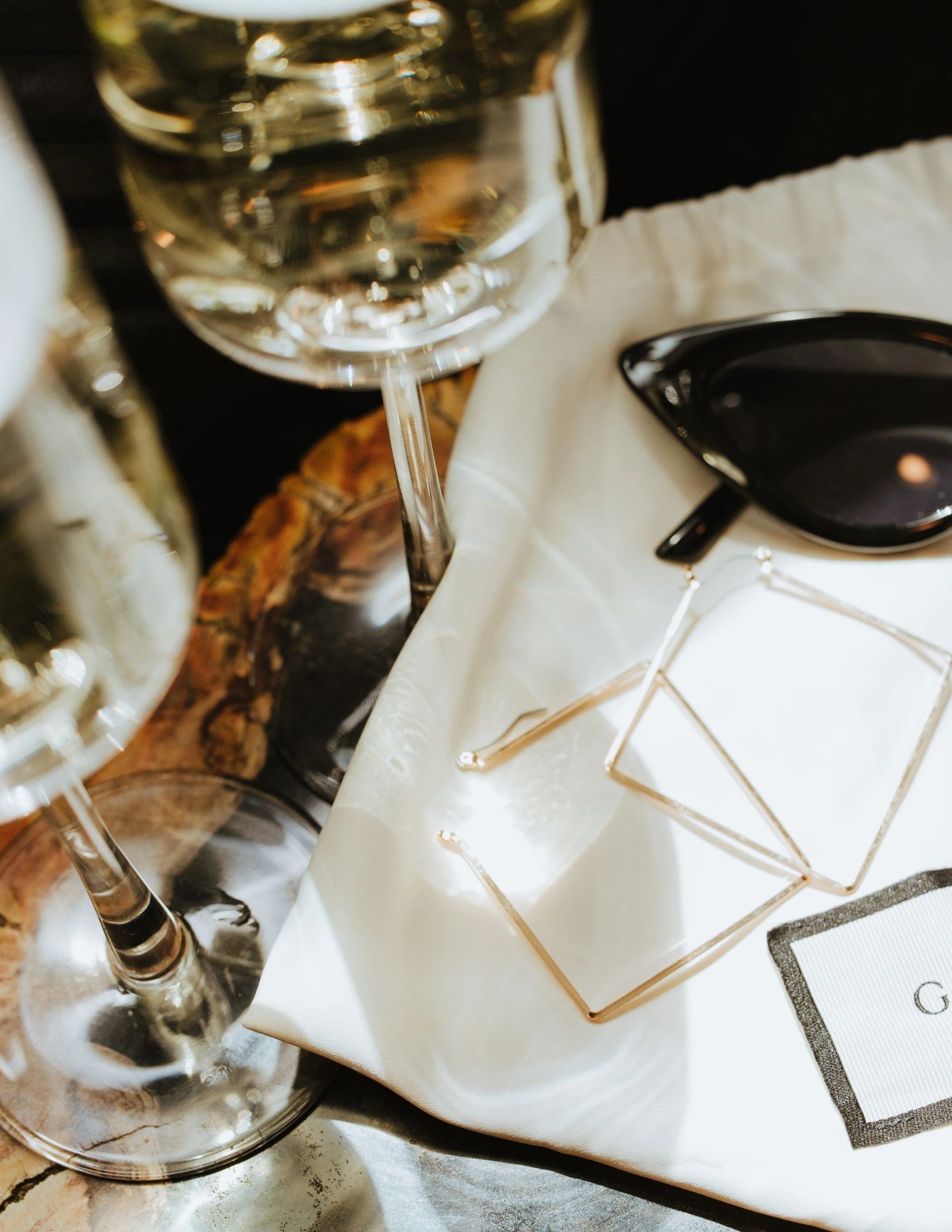 Elegant sunglasses and geometric earrings on a white cloth near wine glasses.