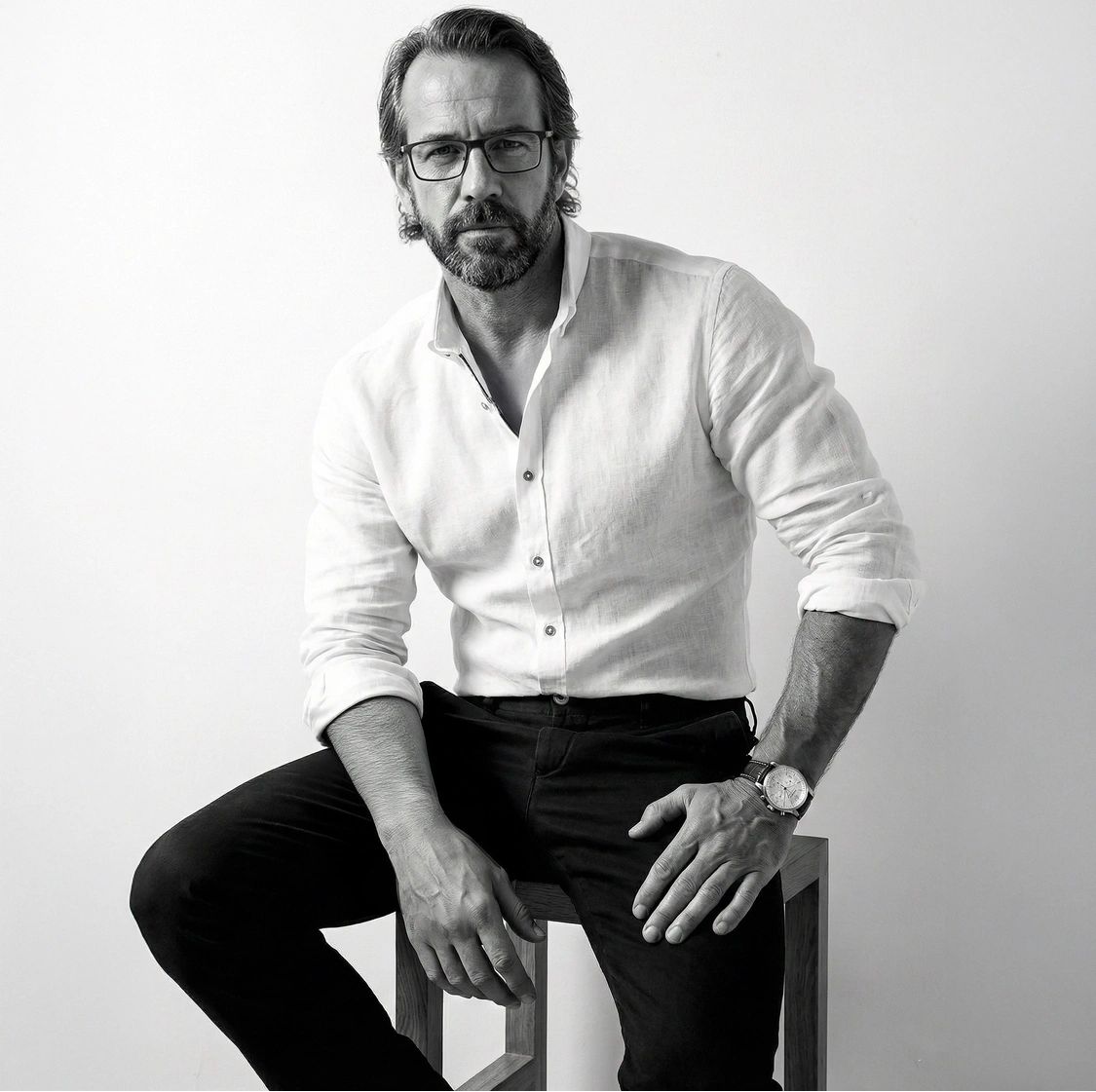Man in glasses and white shirt sitting on a stool, looking serious.