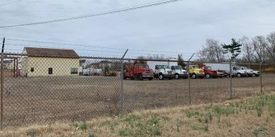 A fenced lot with several parked trucks and a small building under cloudy skies.