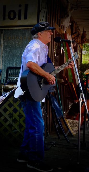 Older man playing guitar and singing into a microphone on a rustic porch.