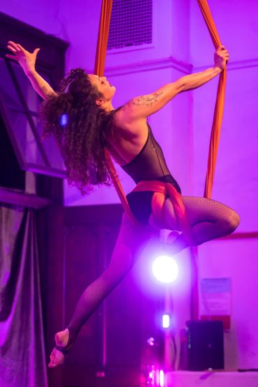 Aerial silk performer gracefully suspended mid-air with dramatic purple lighting.