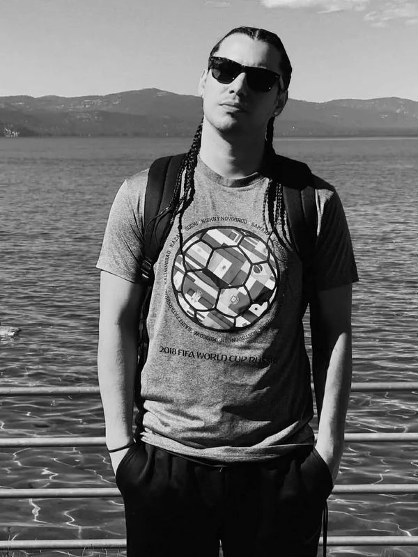 Man with braided hair and sunglasses stands by a lake with mountains in the background.