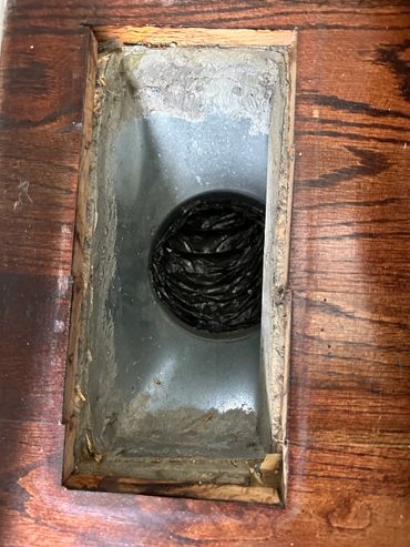 Rectangular opening in wooden floor showing metal duct with black flexible tubing.