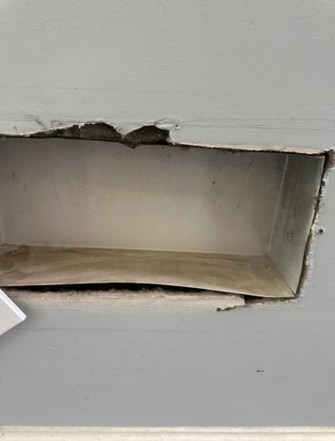 Damaged wall with a rectangular hole and exposed metal duct inside.
