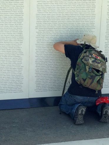 A person kneels reading names on a memorial wall, wearing a camouflage backpack and holding a cap.