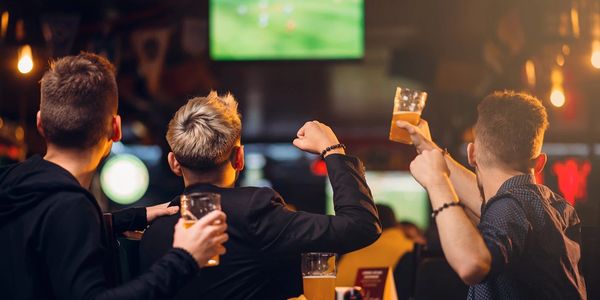 Three men watching a game on TV at a bar, holding drinks and cheering.