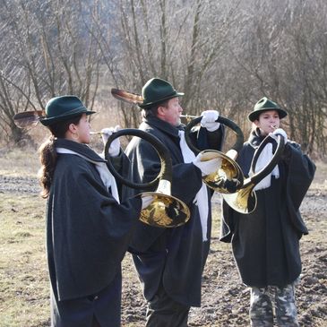 Three musicians in traditional attire playing hunting horns outdoors.