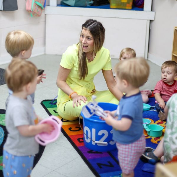 An educator sits on a brightly colored rug surrounded by inquisitive toddlers. STEM learning.