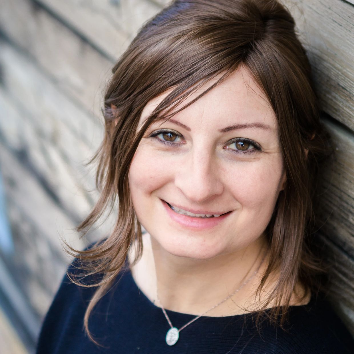 Smiling woman with brown hair leaning against a wooden wall.