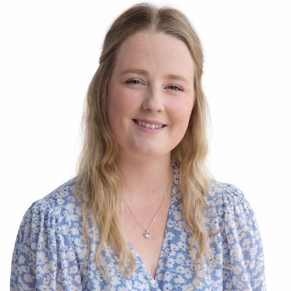 Smiling woman in a blue floral dress with long blonde hair and a pendant necklace.