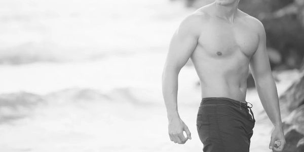 Shirtless man standing by the beach with waves in the background.